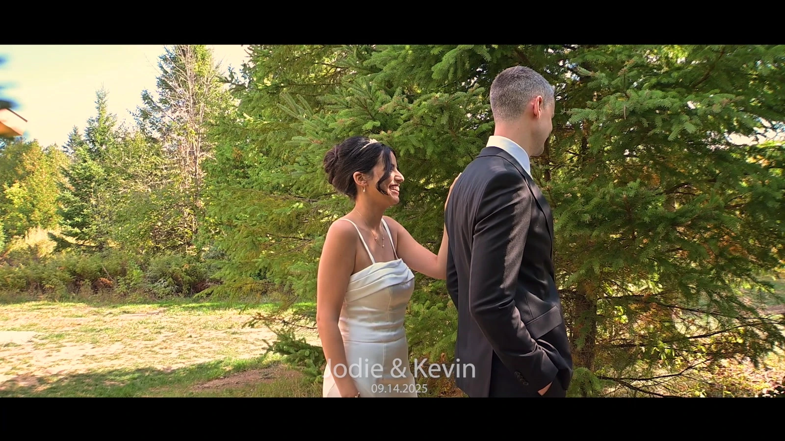 Jodie approaching Kevin from behind beside evergreen trees